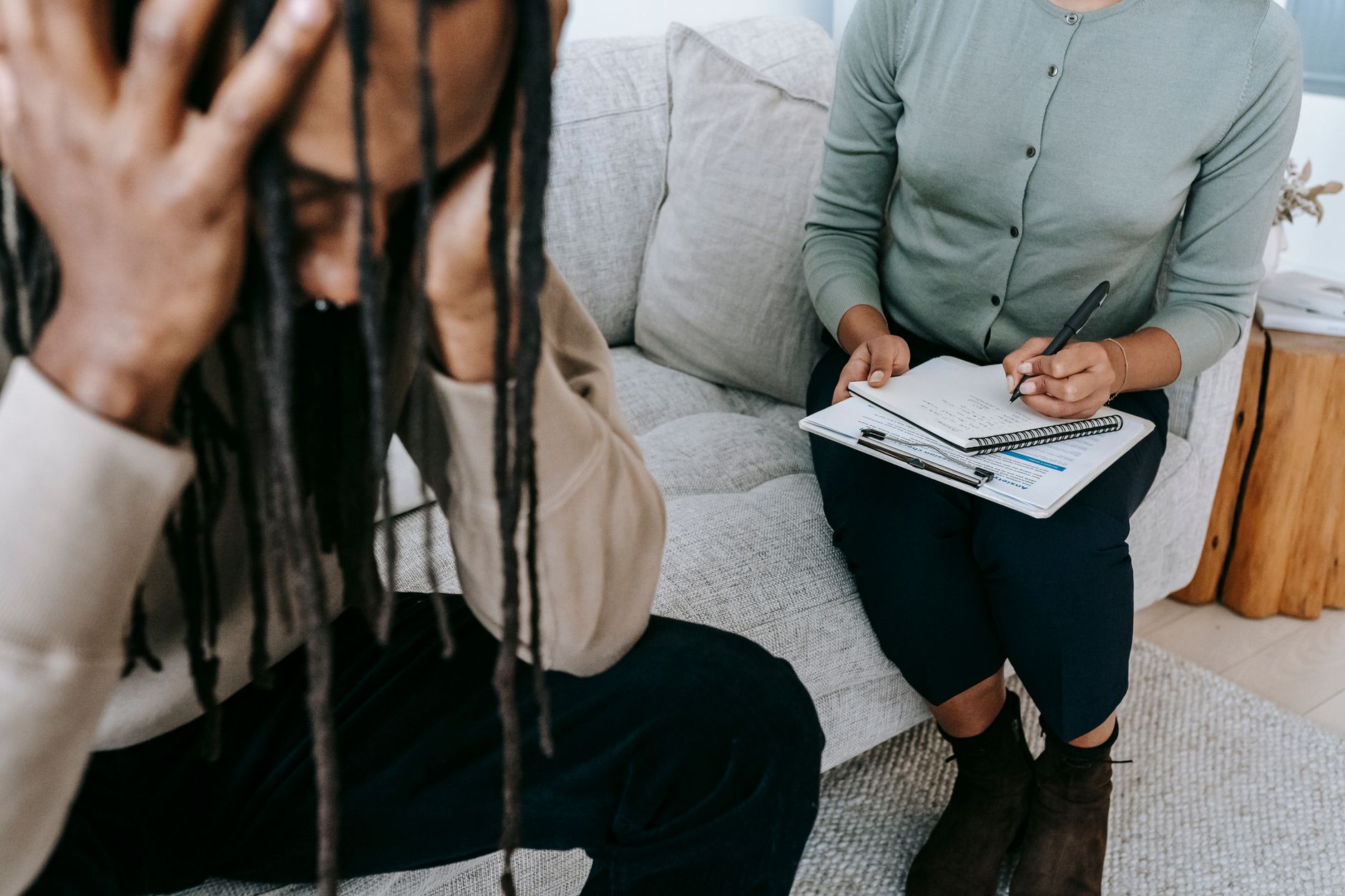 Crop concerned African American male sitting on sofa with female psychologist and holding head in hands in dismay during psychotherapy sessi