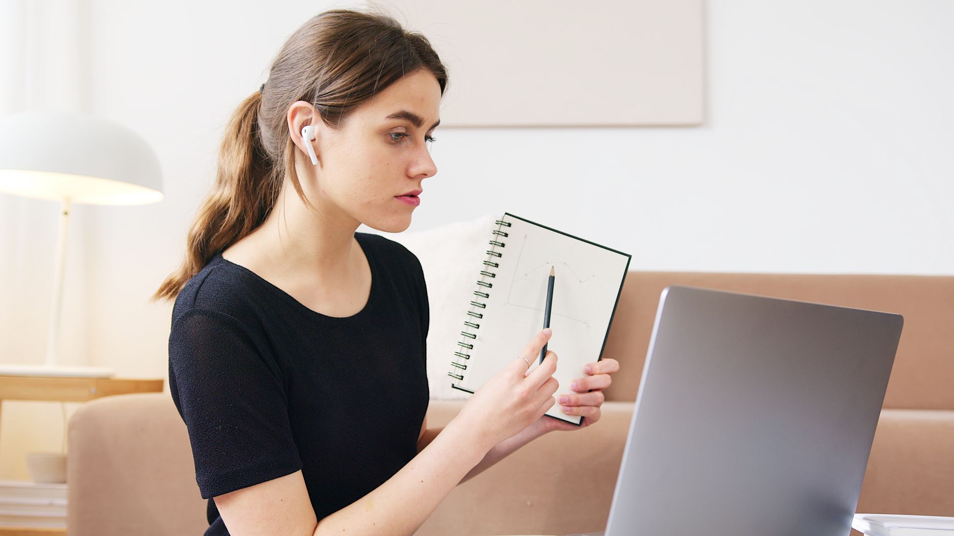 Serious young female student in casual black shirt participating in online webinar via laptop and showing notebook with homework during quar