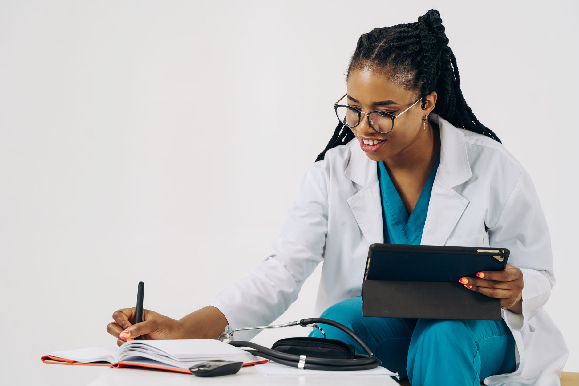 Smiling female doctor in scrubs using tablet and taking notes in a healthcare setting.
