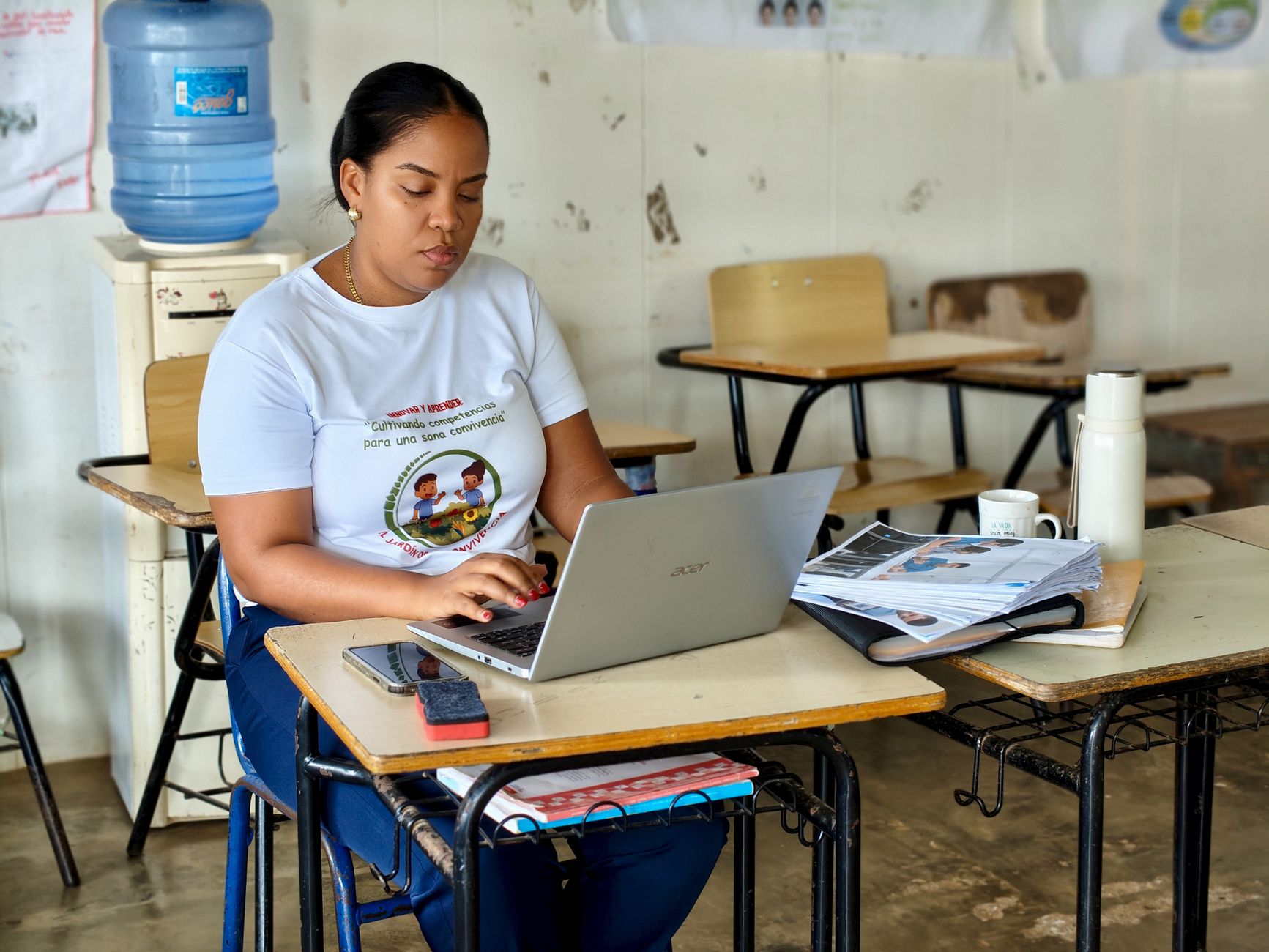 A woman in a classroom typing on a laptop, surrounded by educational materials, conveying a focus on work and study.