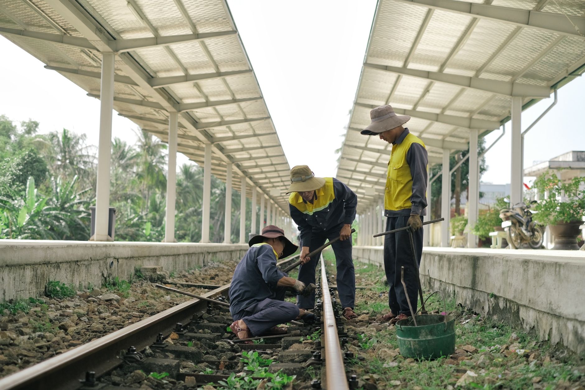 Three workers repairing railway tracks at a rural station during the day.