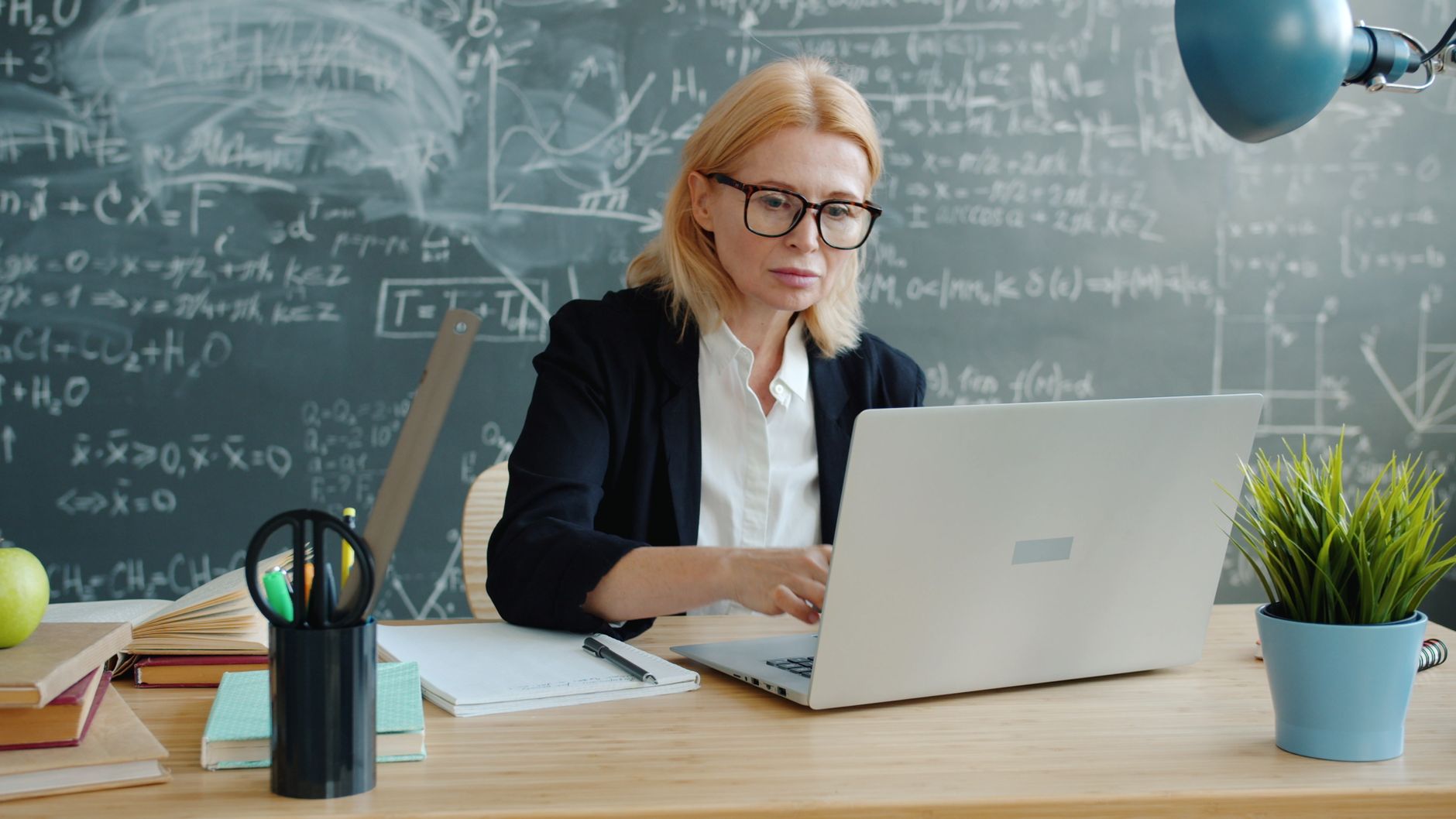 Teacher working on laptop with chalkboard full of equations in background.