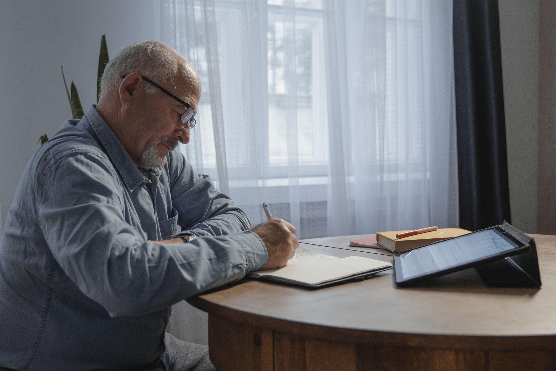 Elderly man engaged in online meeting and writing notes at home office.