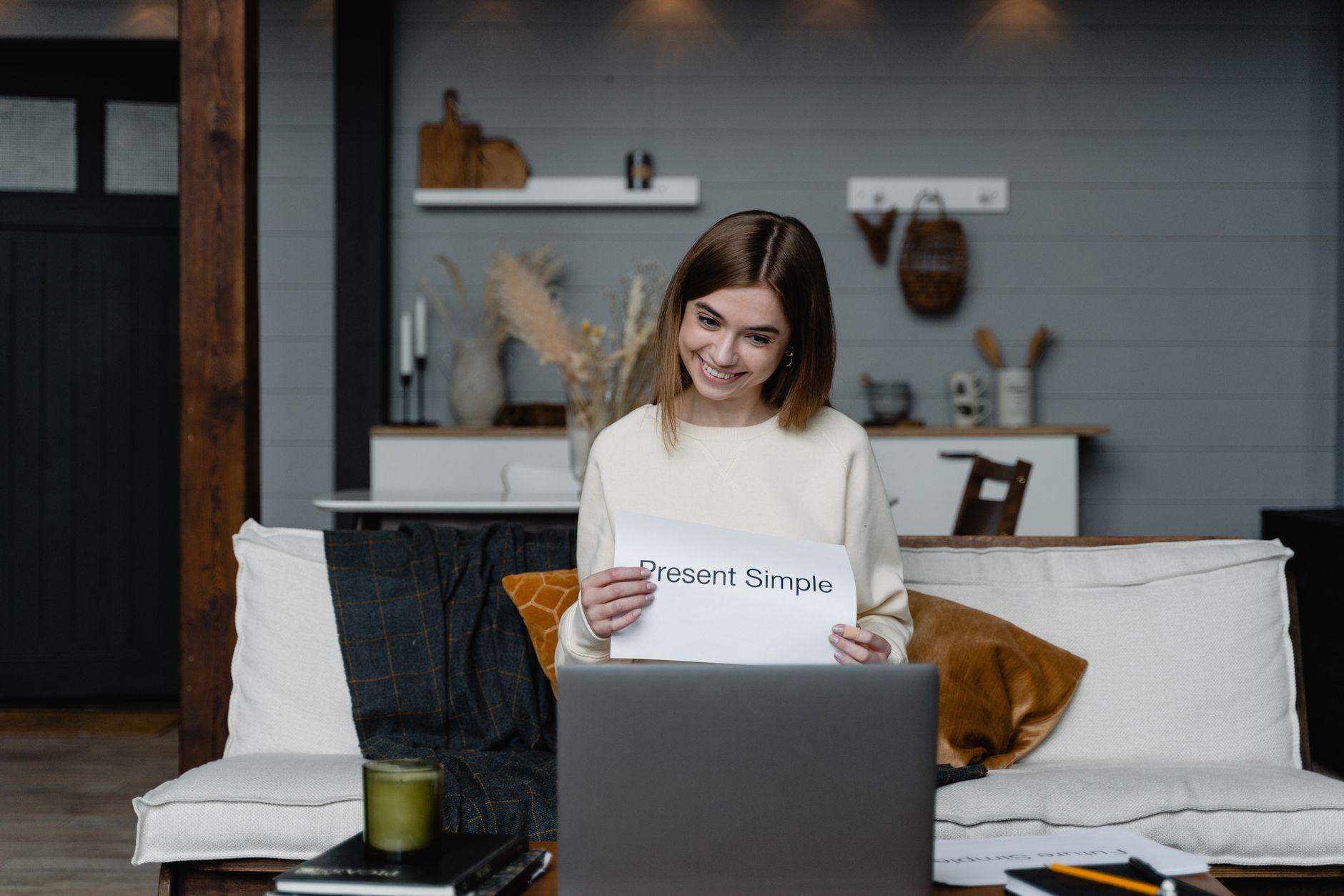 Smiling woman teaching present simple tense from home using a laptop.